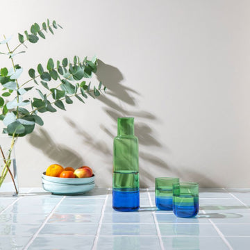 Glaskaraffe „Ocean“ in grün-blauer Multicolor-Optik mit zwei Trinkgläsern auf hellem Tisch; dekoratives Setting mit Schale und Zweigen im Hintergrund.
