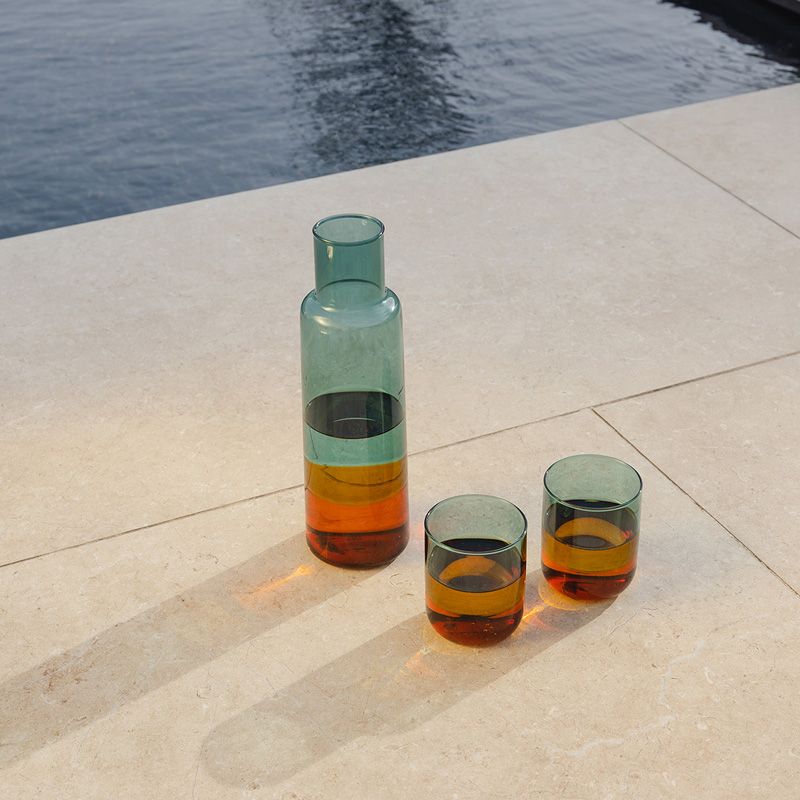 Glaskaraffe in grün-orangefarbener Multicolor-Optik mit zwei Trinkgläsern auf heller Steinfläche neben Wasser; Lichtreflexe betonen das Farbspiel im Glas.
