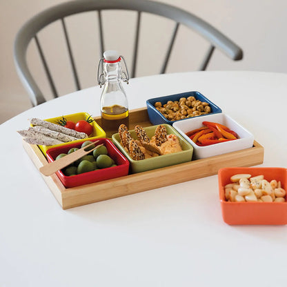 Nahaufnahme des Multicolor-Schalensets auf Holztablett, gefüllt mit Oliven, Dip, Nüssen, Tomaten und Gemüsesticks; Ölflasche im Hintergrund.