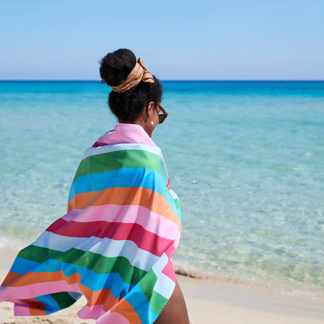 Person trägt ein gestreiftes Strandtuch in Pink, Hellrosa, Mint, Grün, Hellblau und Orange am Strand vor dem Meer
