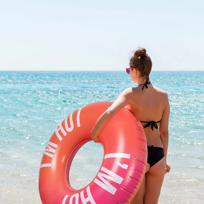 Person von hinten mit dem FISURA Floatie „I’m Hot“ am Ufer mit Blick aufs Meer