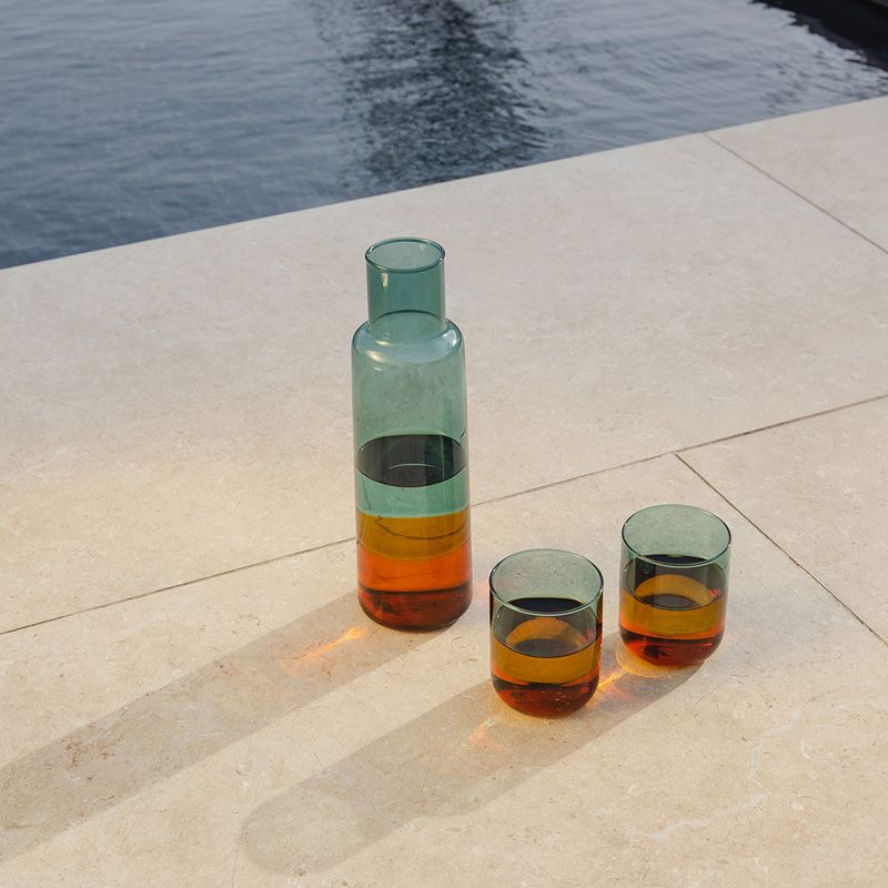 Zwei farbige Trinkgläser aus Borosilikatglas neben passender Glaskaraffe auf heller Steinfläche am Wasser; Lichtreflexe betonen den multicolor Farbverlauf.
