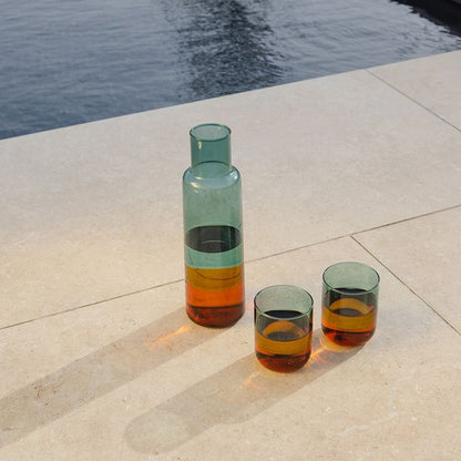 Zwei farbige Trinkgläser aus Borosilikatglas neben passender Glaskaraffe auf heller Steinfläche am Wasser; Lichtreflexe betonen den multicolor Farbverlauf.