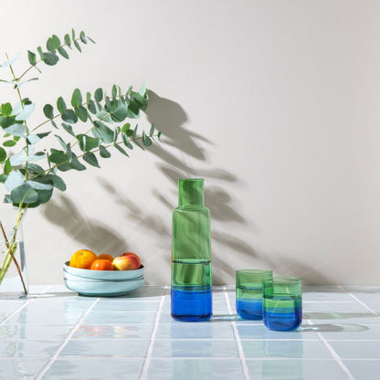 Glaskaraffe „Ocean“ in grün-blauer Multicolor-Optik mit zwei Trinkgläsern auf hellem Tisch; dekoratives Setting mit Schale und Zweigen im Hintergrund.