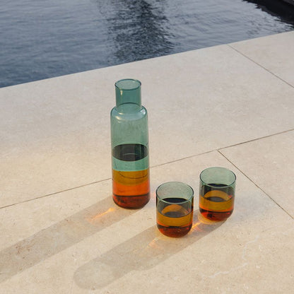 Glaskaraffe in grün-orangefarbener Multicolor-Optik mit zwei Trinkgläsern auf heller Steinfläche neben Wasser; Lichtreflexe betonen das Farbspiel im Glas.