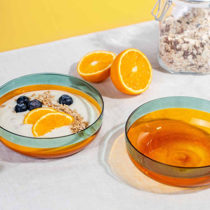 Zwei Glasschälchen „Amber“ auf dem Tisch, eines gefüllt mit Joghurt, Müsli, Blaubeeren und Orange; farbiger Glasverlauf in Türkis und Amber leuchtet im Licht.