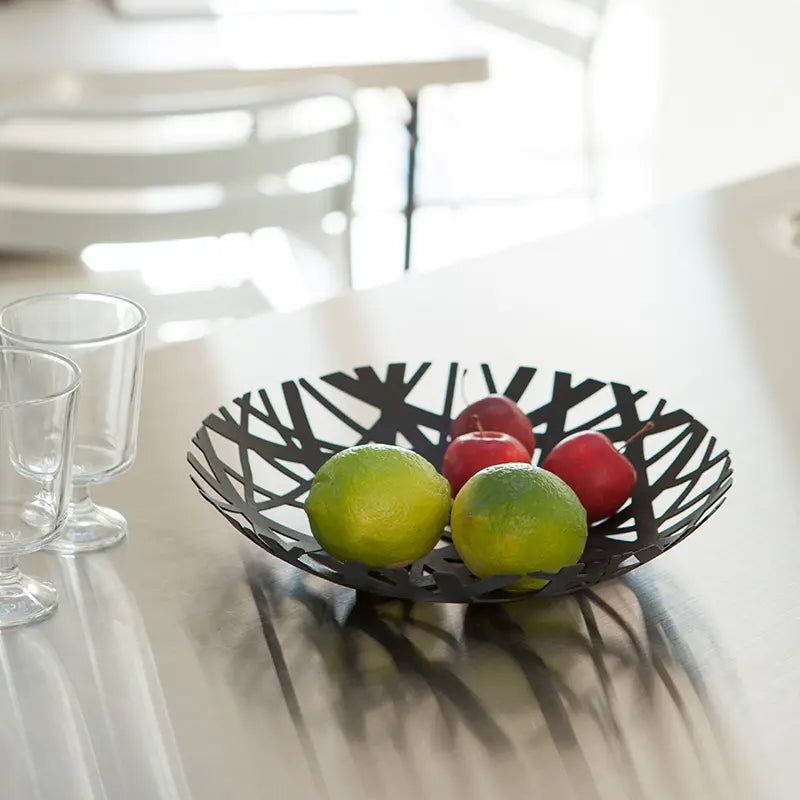Schwarzer Obstkorb mit Nestdesign aus Metall mit Limetten und roten Äpfeln auf einem Tisch.