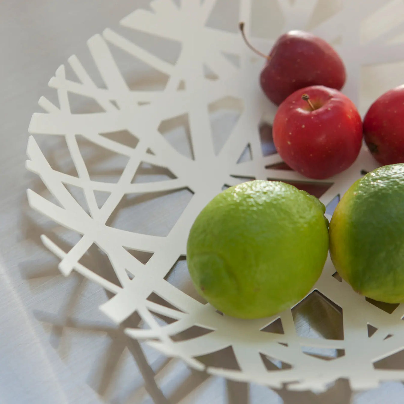 Nahaufnahme des weissen Obstkorbes mit Nestdesign aus Metall mit Limetten und roten Äpfeln.