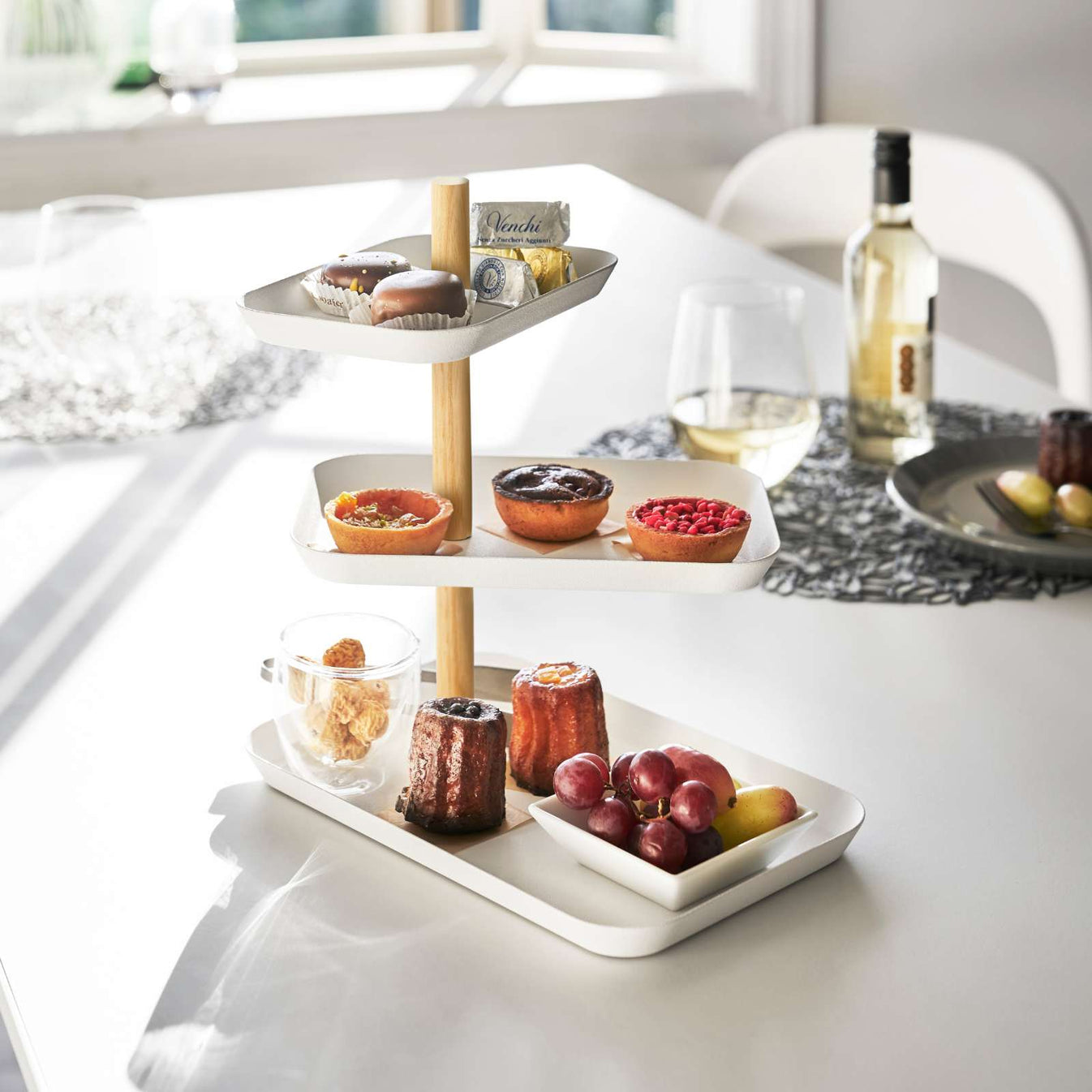 Rechteckige 3-stöckige Étagère in Weiss auf einem gedeckten Tisch, befüllt mit Gebäck, Trauben, Früchten und kleinen Süssigkeiten.