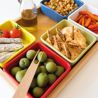 Nahaufnahme des bunten Schalensets mit Holztablett: quadratische Porzellanschalen in Gelb, Rot, Grün, Blau und Weiss, gefüllt mit Oliven, Dip, Nüssen, Tomaten, Gemüsesticks und Brotstangen.