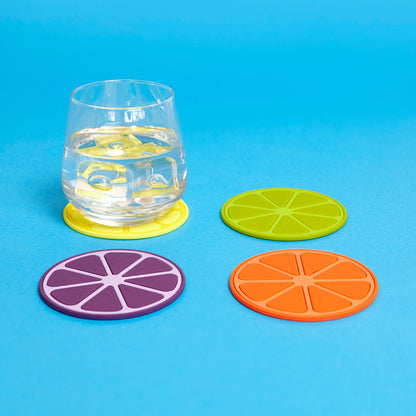 Ein Trinkglas mit Eis steht auf einem gelben fruchtscheibenförmigen Untersetzer. Drei weitere runde Untersetzer in Grün, Lila und Orange liegen daneben auf blauem Hintergrund.