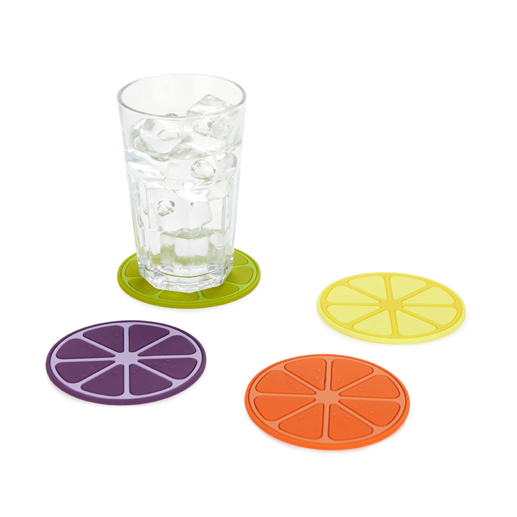 Ein Glas mit Eis steht auf einem grünen runden Untersetzer aus Silikon. Daneben liegen drei weitere Untersetzer in Gelb, Lila und Orange. Alle Untersetzer sind wie Fruchtscheiben gestaltet.