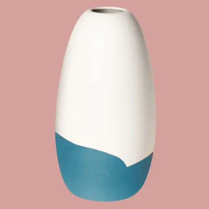 Konische Porzellanvase „Iapetus - petrol“, seidenmatt weiss glasiert mit petrol Akzent am unteren Bereich, freigestellt vor hellrotem Hintergrund.