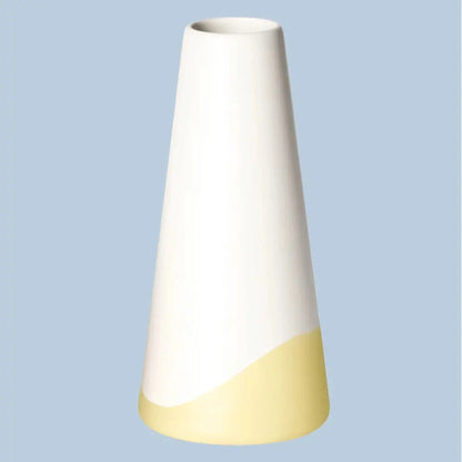 Schlanke, konische Porzellanvase „Neptun“ in seidenmattem Weiss mit gelbem Farbschwung am Fuss, freigestellt vor hellblauem Hintergrund.