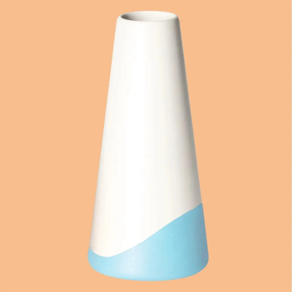 Konische Vase „Neptun“ aus Porzellan, weiss seidenmatt glasiert mit hellblauem Farbakzent am unteren Rand, freigestellt vor apricotfarbenem Hintergrund.