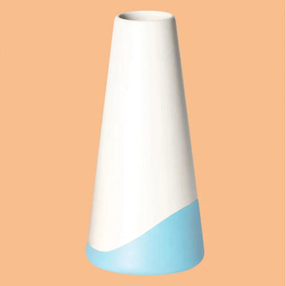Konische Vase „Neptun“ aus Porzellan, weiss seidenmatt glasiert mit hellblauem Farbakzent am unteren Rand, freigestellt vor apricotfarbenem Hintergrund.