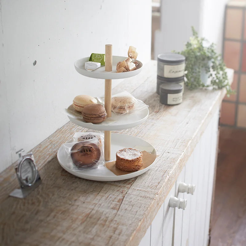 Weisse dreistöckige Etagère mit hellem Holzgestänge auf einem Holztisch, befüllt mit Macarons, Gebäck und kleinen Süssigkeiten in einem hellen Innenraum.