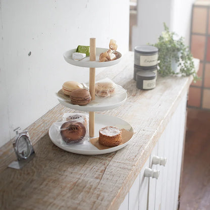 Weisse dreistöckige Etagère mit hellem Holzgestänge auf einem Holztisch, befüllt mit Macarons, Gebäck und kleinen Süssigkeiten in einem hellen Innenraum.